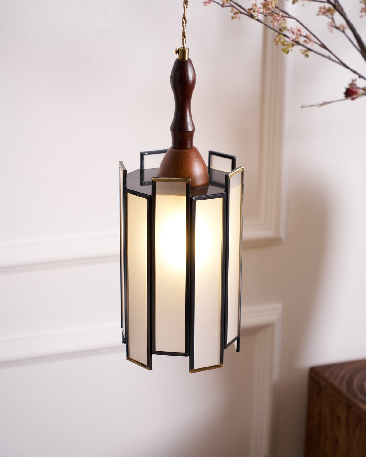 Lit Clérachic frosted-panel pendant against a panelled wall, with a wooden plinth and blossom branches at the edges.
