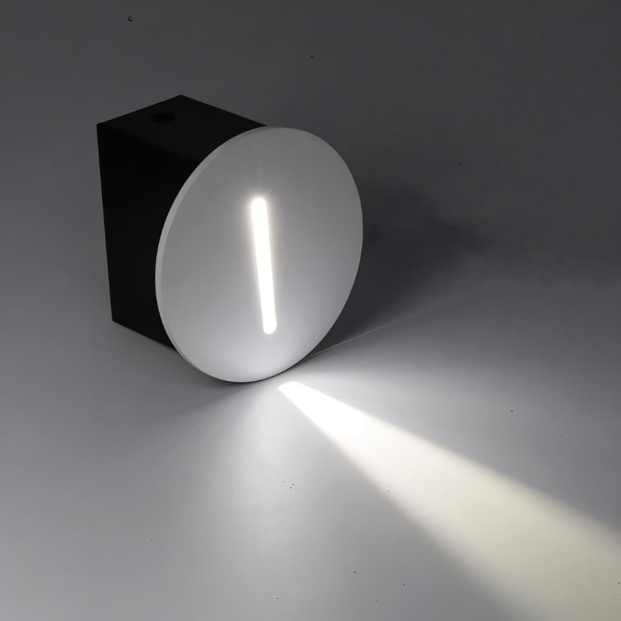 Clérachic round stair light with a white face plate shown at an angle on its black housing box, the slot lit cool white and a bright shaft crossing the grey surface.