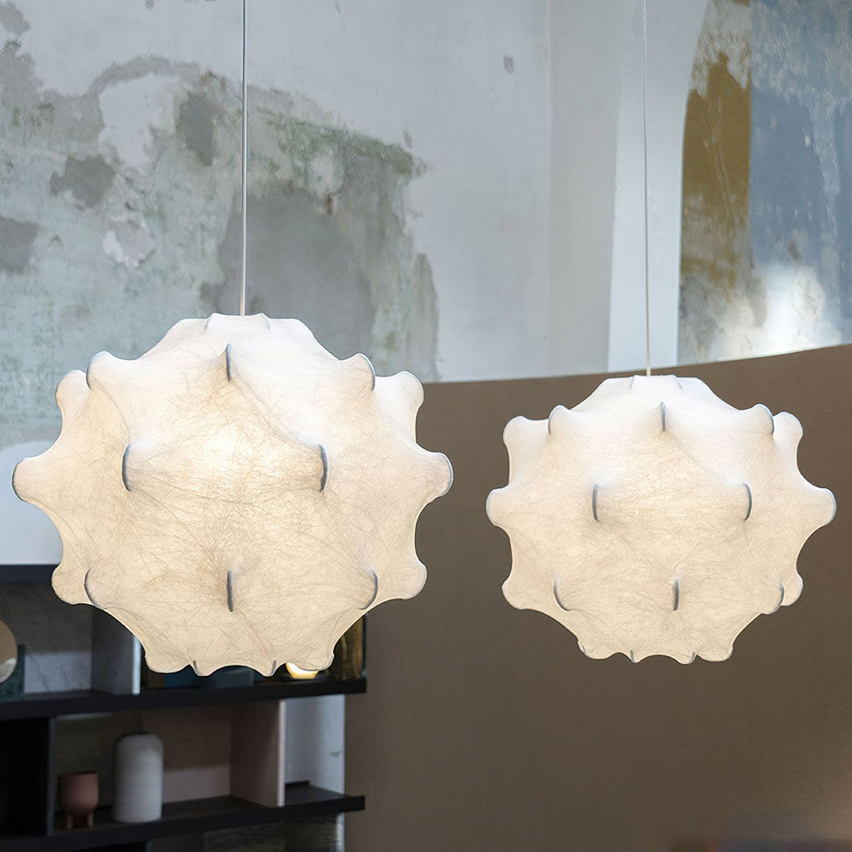 Two white Clérachic pendants of different sizes hang side by side, glowing against a weathered plaster wall and a dark shelf.