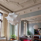 A white Clérachic pendant hangs in a tall room with ornate cornices, with a round paper lantern and a sofa behind.