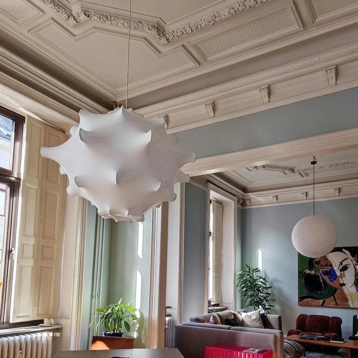 A white Clérachic pendant hangs in a tall room with ornate cornices, with a round paper lantern and a sofa behind.