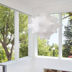 A white Clérachic pendant hangs by a corner of tall windows, with green trees and a bright sky outside.