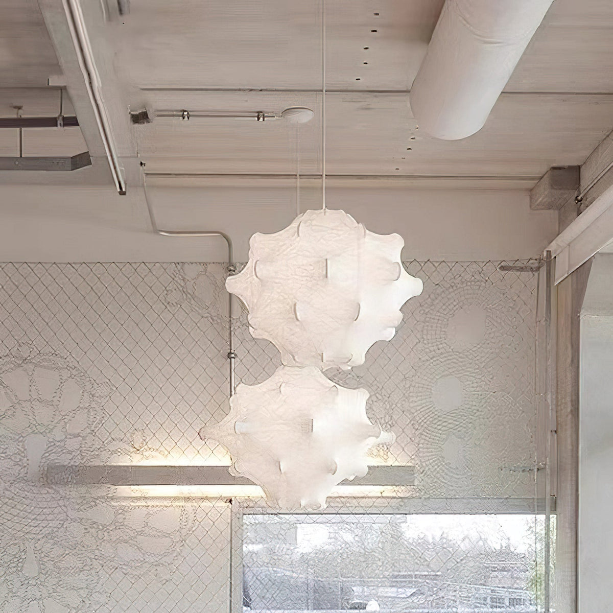 Two white Clérachic pendants hang one above the other in a pale loft with exposed ducting and a lace-patterned wall.