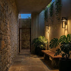 Two Clérachic steel wall lights flank a glazed door in a sunlit courtyard with terracotta chairs and greenery.