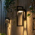 At dusk, two Clérachic steel wall lights cast a warm glow beside a glazed door in the same paved courtyard.