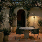 A Clérachic steel wall light warms a pergola-covered patio with a mosaic dining table set for evening.