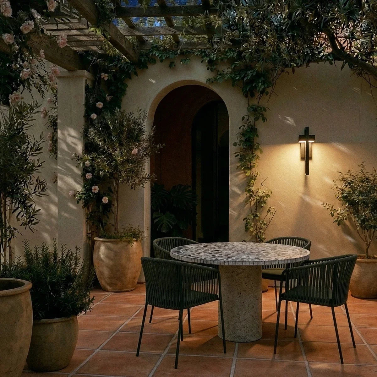A Clérachic steel wall light warms a pergola-covered patio with a mosaic dining table set for evening.
