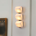 A lit Clérachic wall light of three stacked alabaster blocks on a white panelled wall, beside a dark-framed picture.