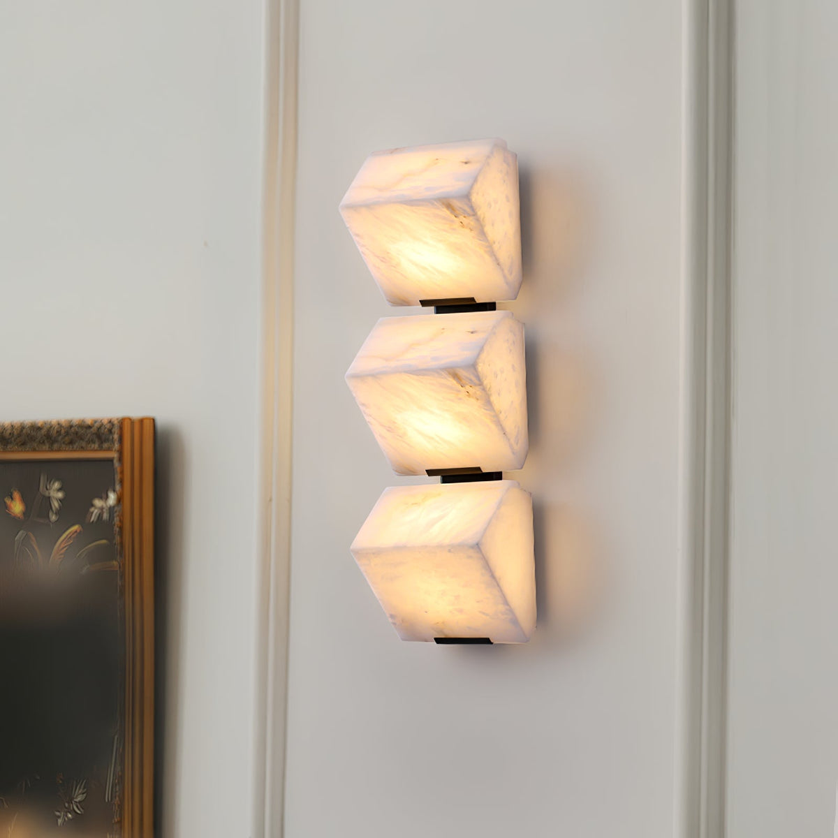 A lit Clérachic wall light of three stacked alabaster blocks on a white panelled wall, beside a dark-framed picture.