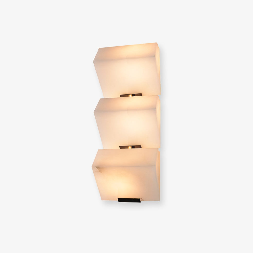 Front view of this Clérachic wall light on a plain white backdrop, three stepped alabaster blocks glowing warm above black wall plates.