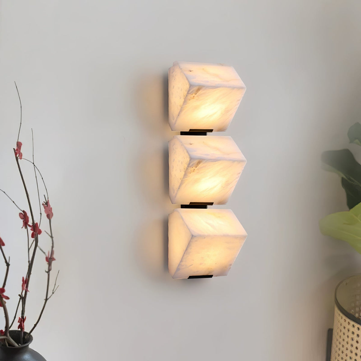 Three stacked alabaster blocks of this Clérachic wall light glow warm on a pale wall, beside bare branches with small red buds in a dark vase.