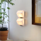 A two-block Clérachic alabaster wall light glows on a pale wall between a leafy potted tree and a dark wooden mirror frame.