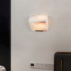 A single Clérachic alabaster wall light with rose-brown veining glows on a pale wall beside a dark door frame and a wall switch panel.