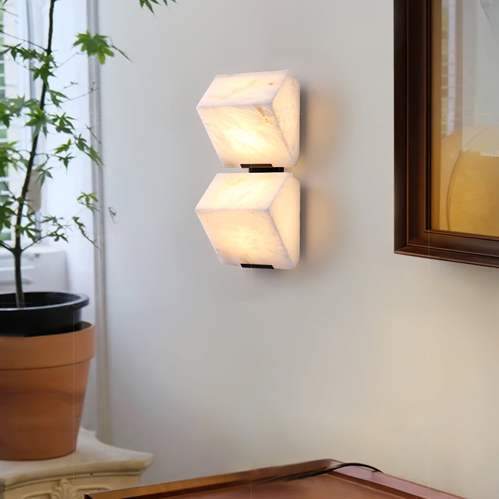 A two-block Clérachic alabaster wall light on a pale wall, with a potted tree to the left and a dark wooden frame above a console.