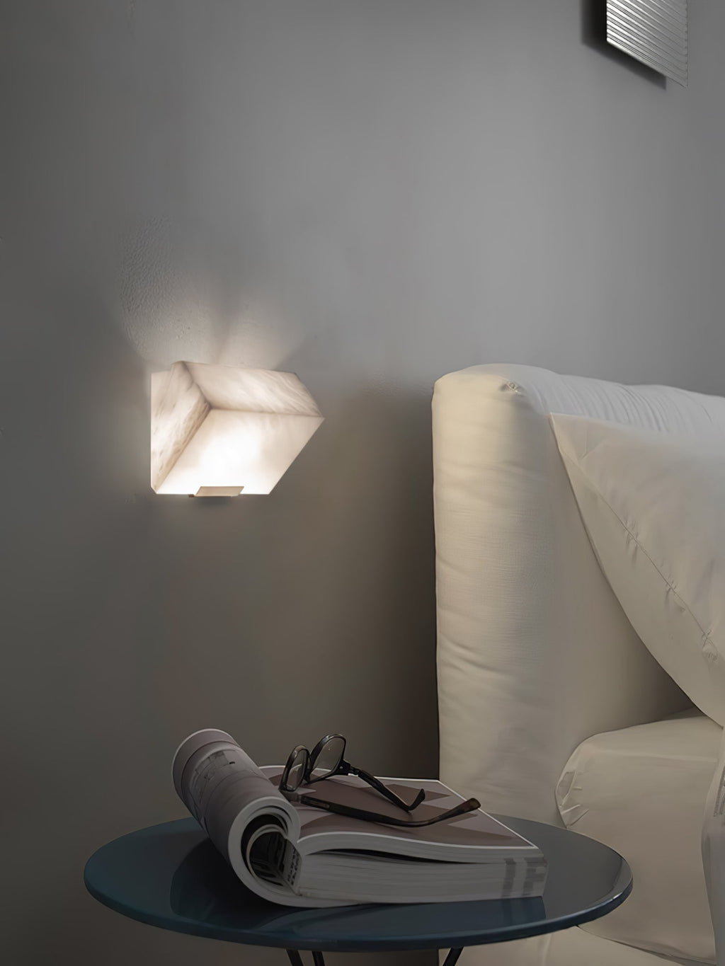 A single Clérachic alabaster wall light glows on a grey wall above a round glass side table with an open magazine and glasses beside a cream sofa.