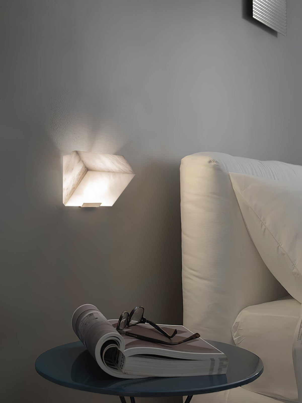 A single Clérachic alabaster wall light glows on a grey wall above a round glass side table with an open magazine and glasses beside a cream sofa.