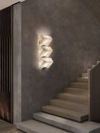 A three-block Clérachic alabaster wall light glows on a textured grey wall beside a floating timber staircase.