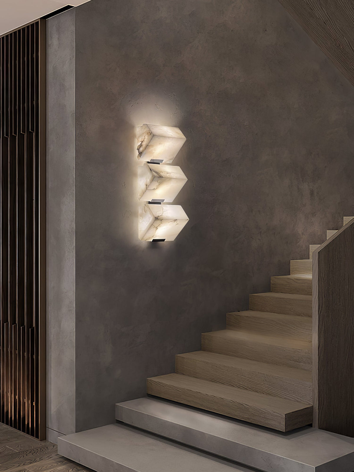 A three-block Clérachic alabaster wall light glows on a textured grey wall beside a floating timber staircase.