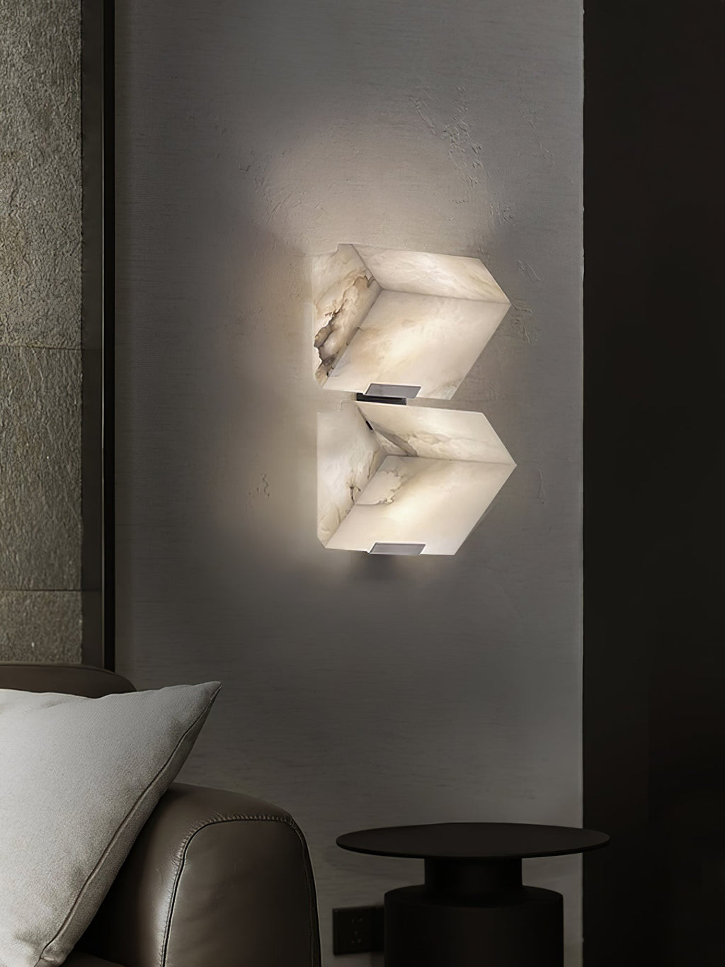 A two-block Clérachic alabaster wall light glows on a dark grey wall above a leather armchair, a pale cushion and a small black side table.