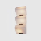 Cut-out view of a three-block Clérachic alabaster wall light on a pale grey backdrop, each block glowing warm above its black plate.