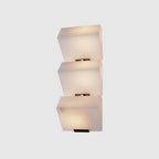 A three-block Clérachic alabaster wall light on a grey backdrop, the stepped stone blocks glowing evenly above black plates.