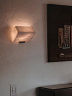 A single Clérachic alabaster wall light glows on a textured plaster wall beside a dark framed artwork and a wall switch.