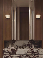 A Clérachic alabaster wall light on each side of a tall mirror above a dark marble vanity in a walnut-panelled bathroom.