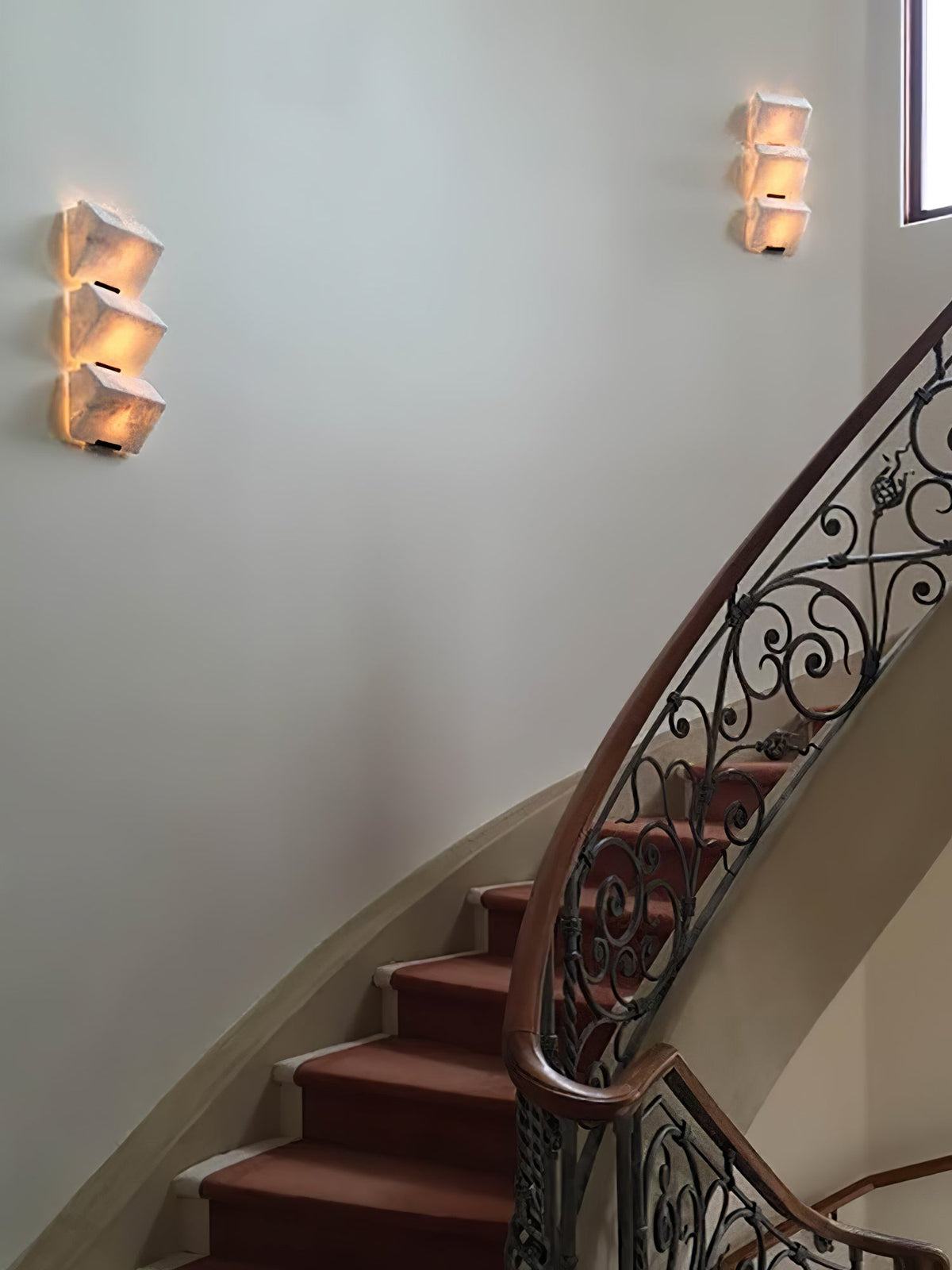 Two Clérachic alabaster wall lights glow on a pale stairwell wall above a curved staircase with an ornate iron balustrade.