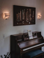 Two Clérachic alabaster wall lights glow on either side of a large framed artwork above a dark upright piano.