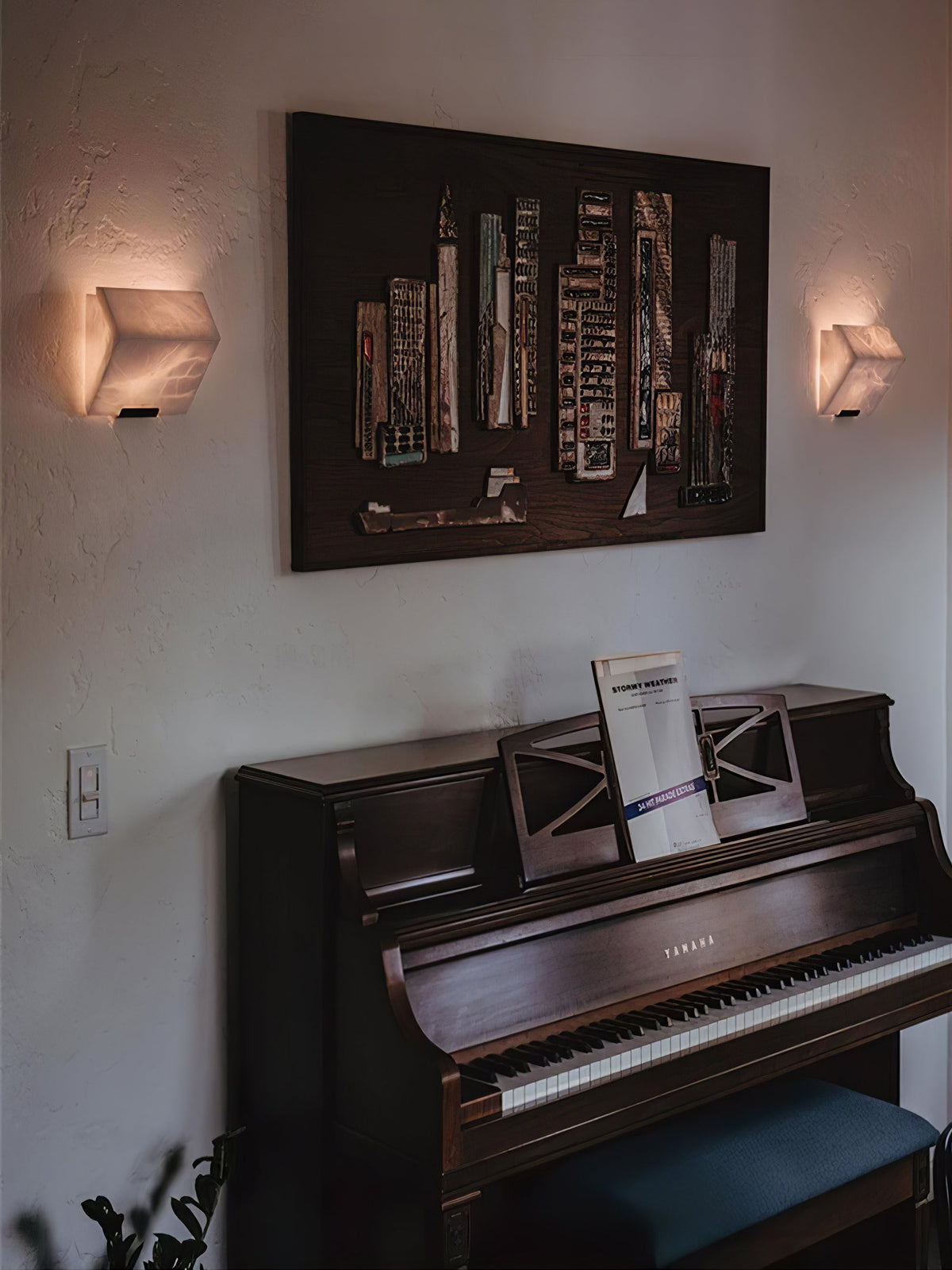 Two Clérachic alabaster wall lights glow on either side of a large framed artwork above a dark upright piano.
