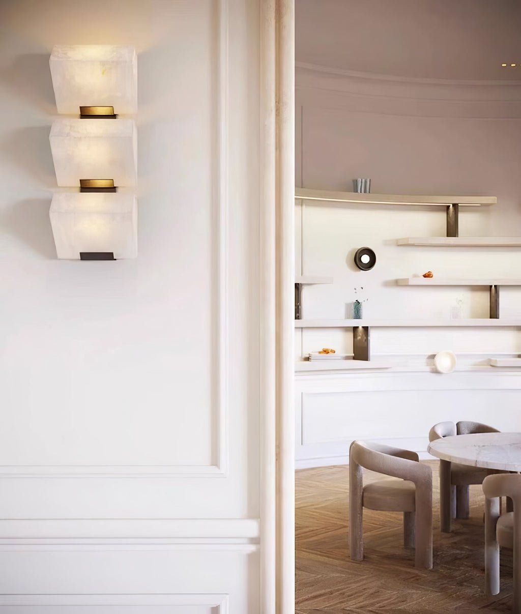 A three-block Clérachic alabaster wall light glows beside a cream doorway, with a dining room and round marble table beyond.