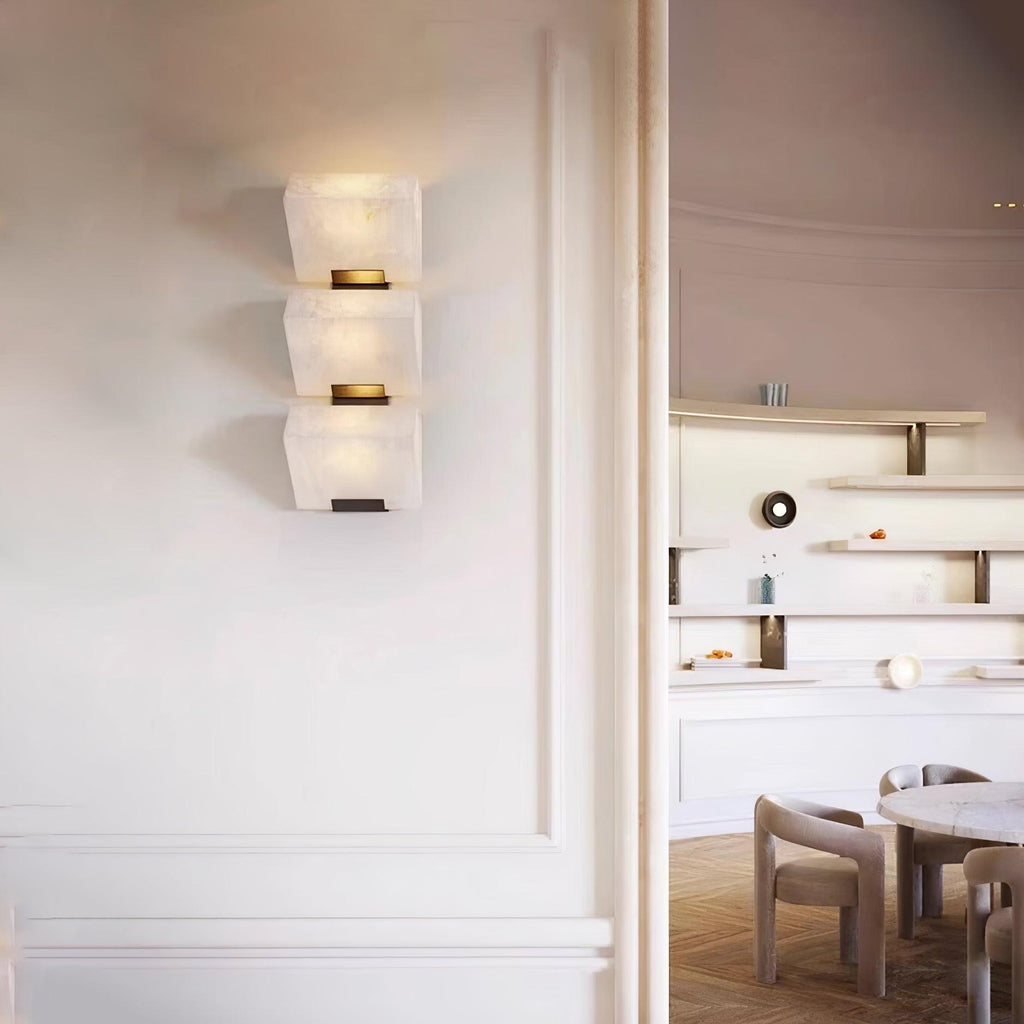 A closer view of the three-block Clérachic alabaster wall light on a white panelled wall beside an open doorway to a dining room.