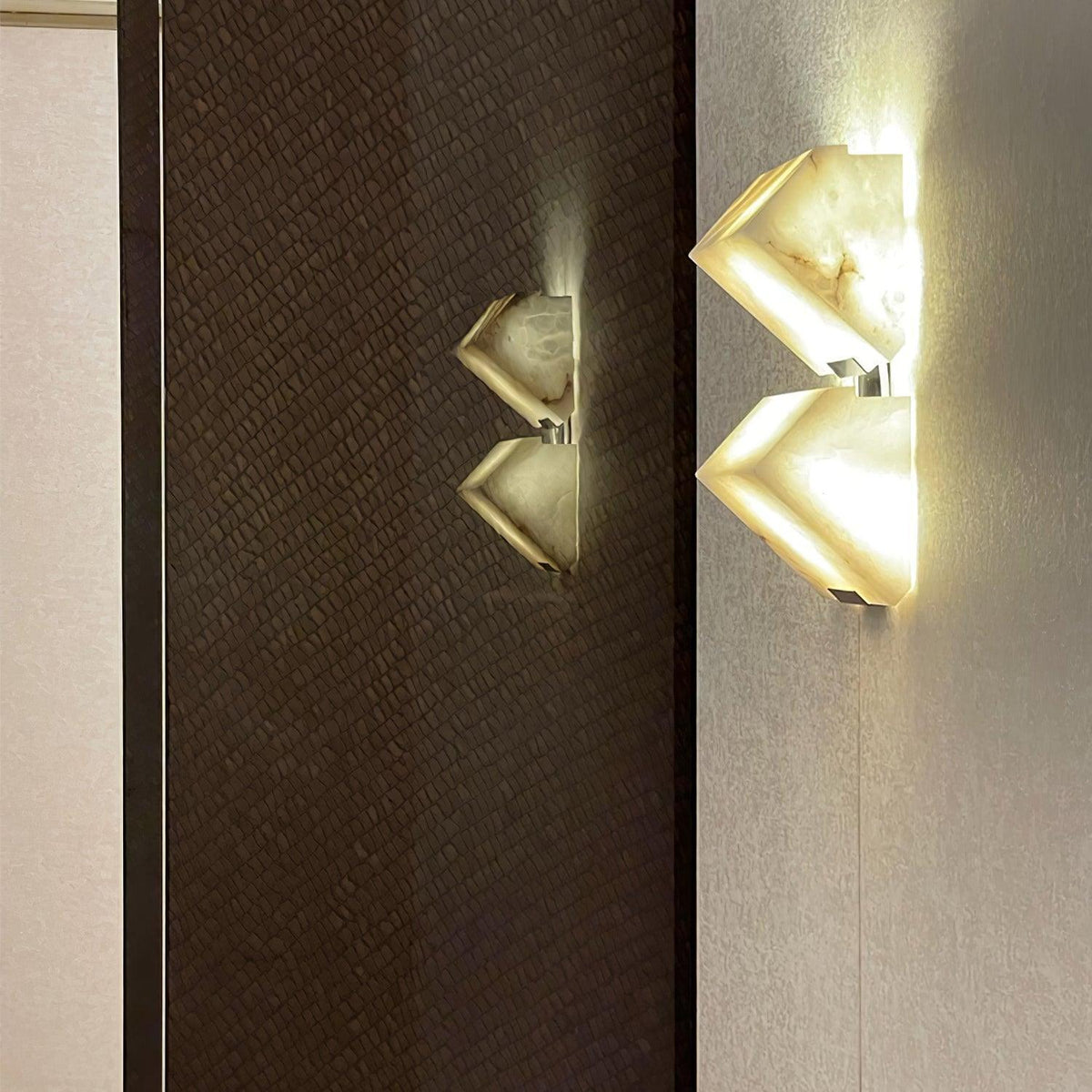 Two Clérachic alabaster wall lights glow on a dark woven wall panel and a pale wall, the brighter one showing the stepped blocks clearly.