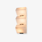 A three-block Clérachic alabaster wall light on a pale backdrop, the stepped stone glowing warm above a slim black plate.