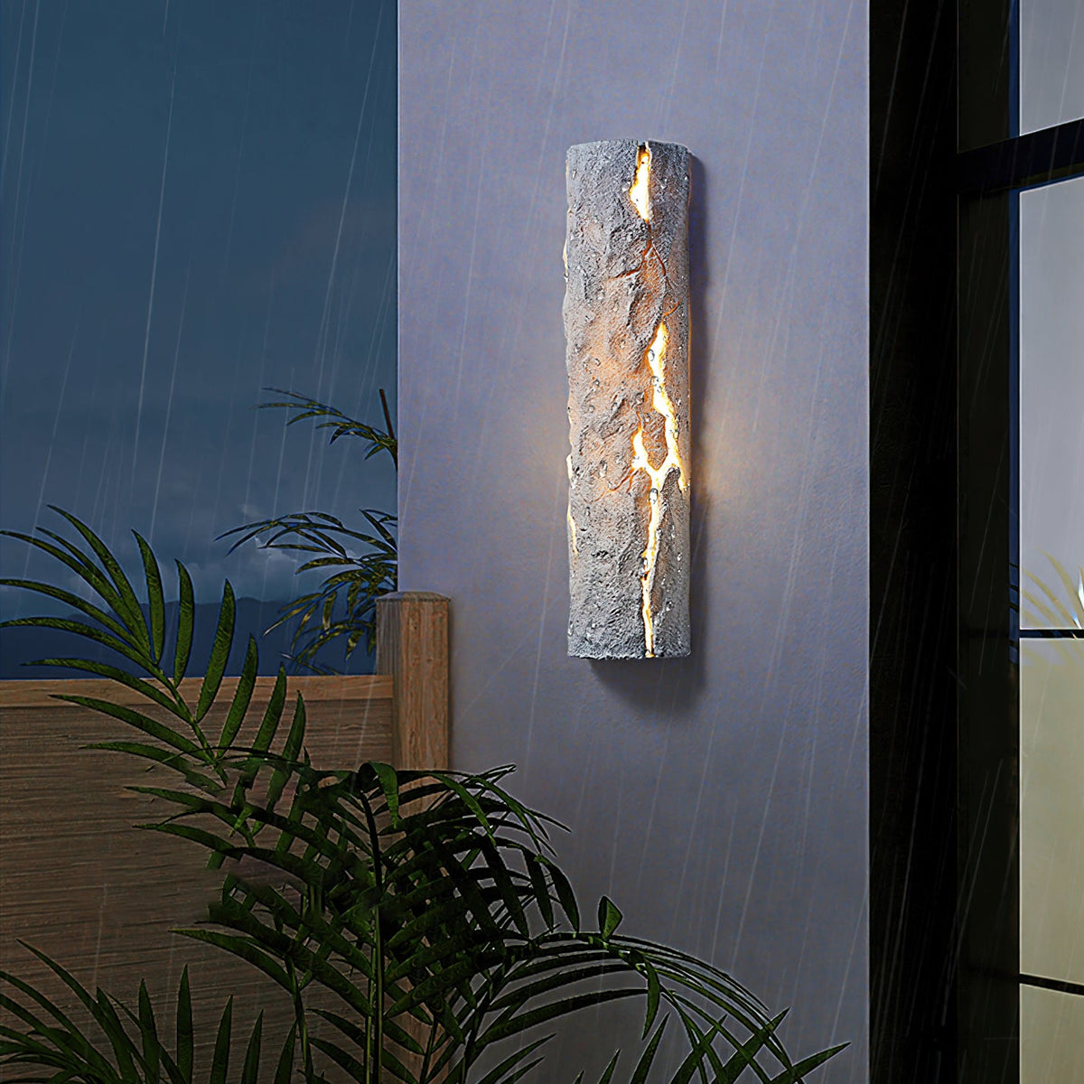 Grey stone-look wall light lit through its cracks on an exterior wall at night, with rain falling and palm leaves nearby, a Clérachic wall light.