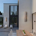 Two grey stone-look wall lights on a pale terrace wall beside tall glass doors, with an outdoor dining table below, from Clérachic.