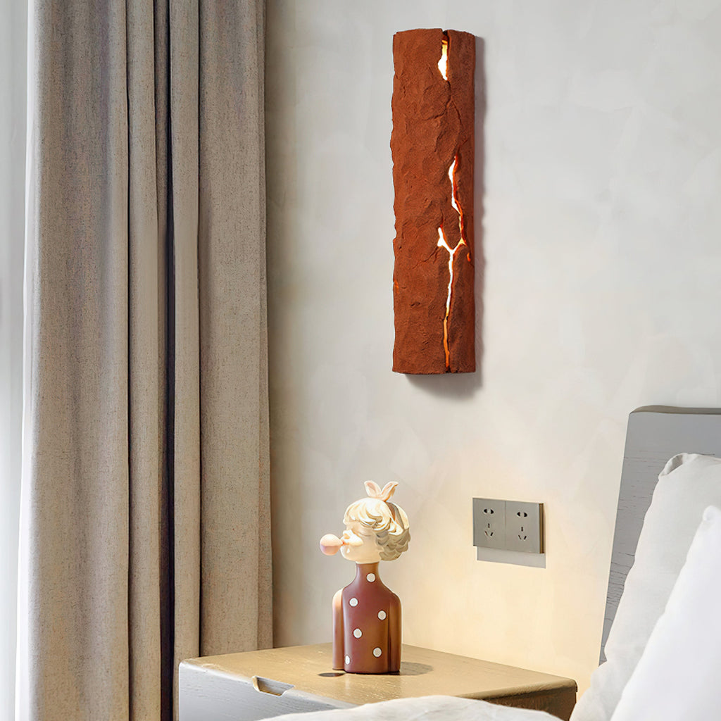 Red stone-look wall light above a bedside table with a small figurine, a grey curtain at its side, a Clérachic wall light.