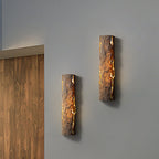 A pair of dark brown stone-look wall lights glowing warm on a grey wall next to a wood-slat panel, from Clérachic.