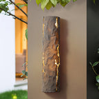 Close view of a dark brown rough-textured wall light with bright cracks, mounted on a taupe wall by leaves, a Clérachic wall light.