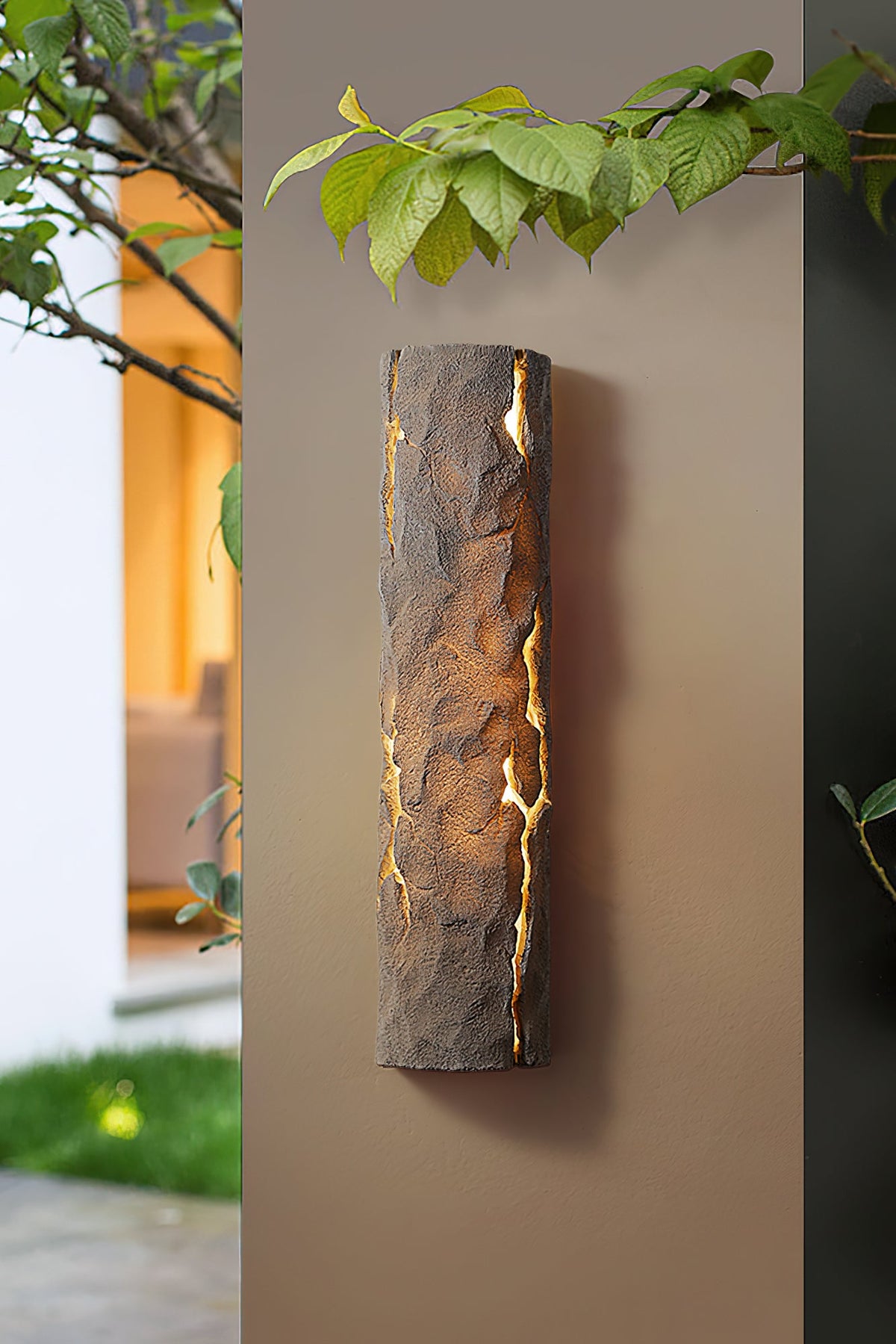 Brown stone-look wall light on a taupe wall under green leaves, with a warmly lit window behind it, from Clérachic.