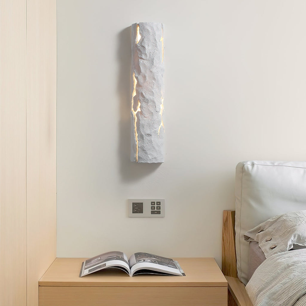 Grey stone-look wall light above a bedside table with an open magazine, near a light wood headboard, from Clérachic.