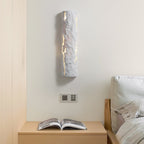 Grey stone-look wall light above a bedside table with an open magazine, near a light wood headboard, from Clérachic.