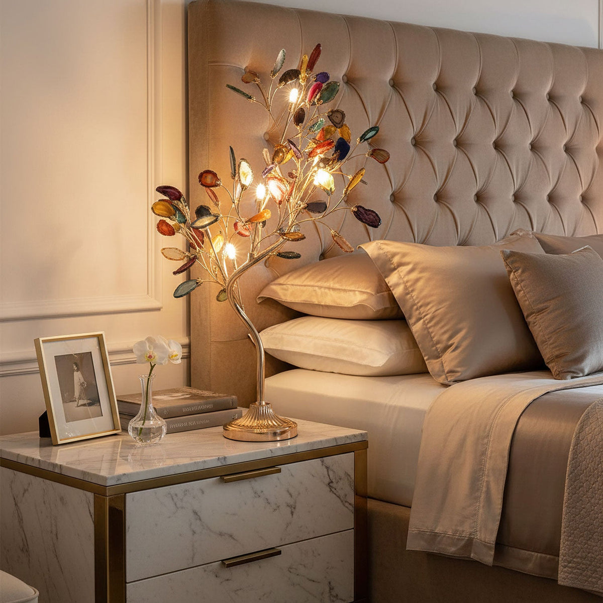 A Clérachic tree-shaped table lamp glows on a marble-topped bedside table, with a tufted headboard and framed photo behind.