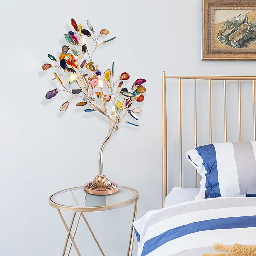 A Clérachic agate-leaf table lamp on a glass-topped side table, beside a brass-framed bed with blue striped pillows.