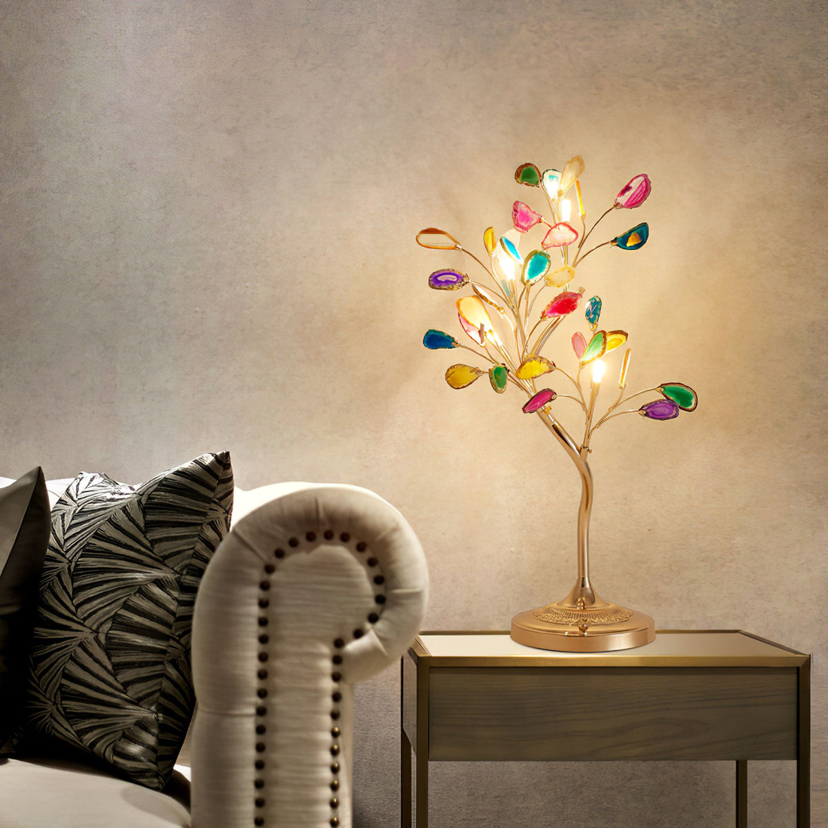 A Clérachic table lamp with glowing multicolour agate leaves stands on a small wooden table next to a cream studded sofa.