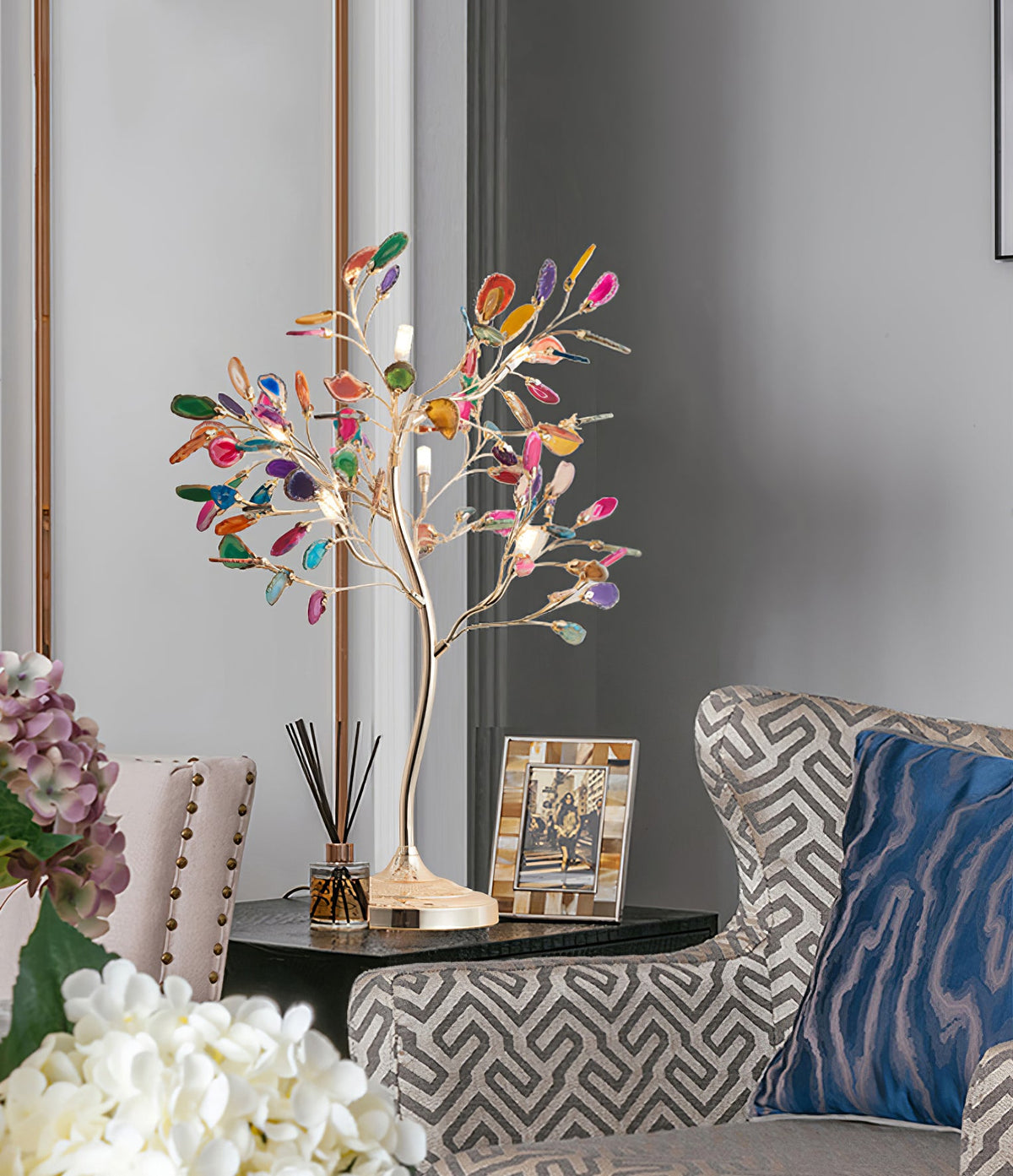 A Clérachic agate-leaf table lamp on a dark side table among a patterned armchair, white flowers and a reed diffuser.