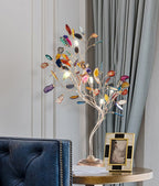 This Clérachic table lamp stands on a round brass side table in front of grey curtains, with a framed photo at its base.