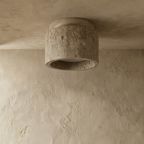 Clérachic compact drum ceiling light switched off, pitted travertine body with a pale wood canopy on a plaster ceiling.