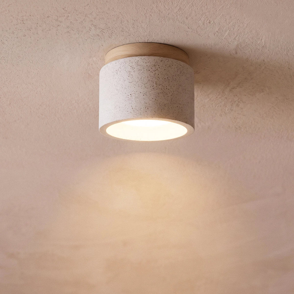 Clérachic compact drum ceiling light in speckled white cement with a wood canopy, glowing softly on a pink plaster ceiling.