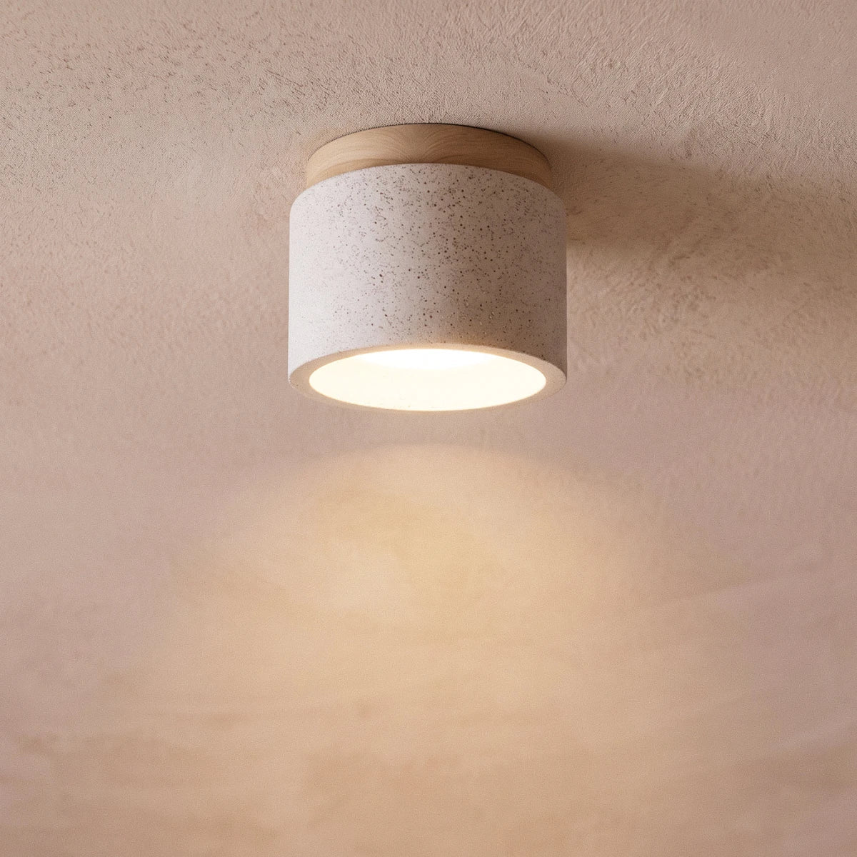 Clérachic compact drum ceiling light in speckled white cement with a wood canopy, glowing softly on a pink plaster ceiling.